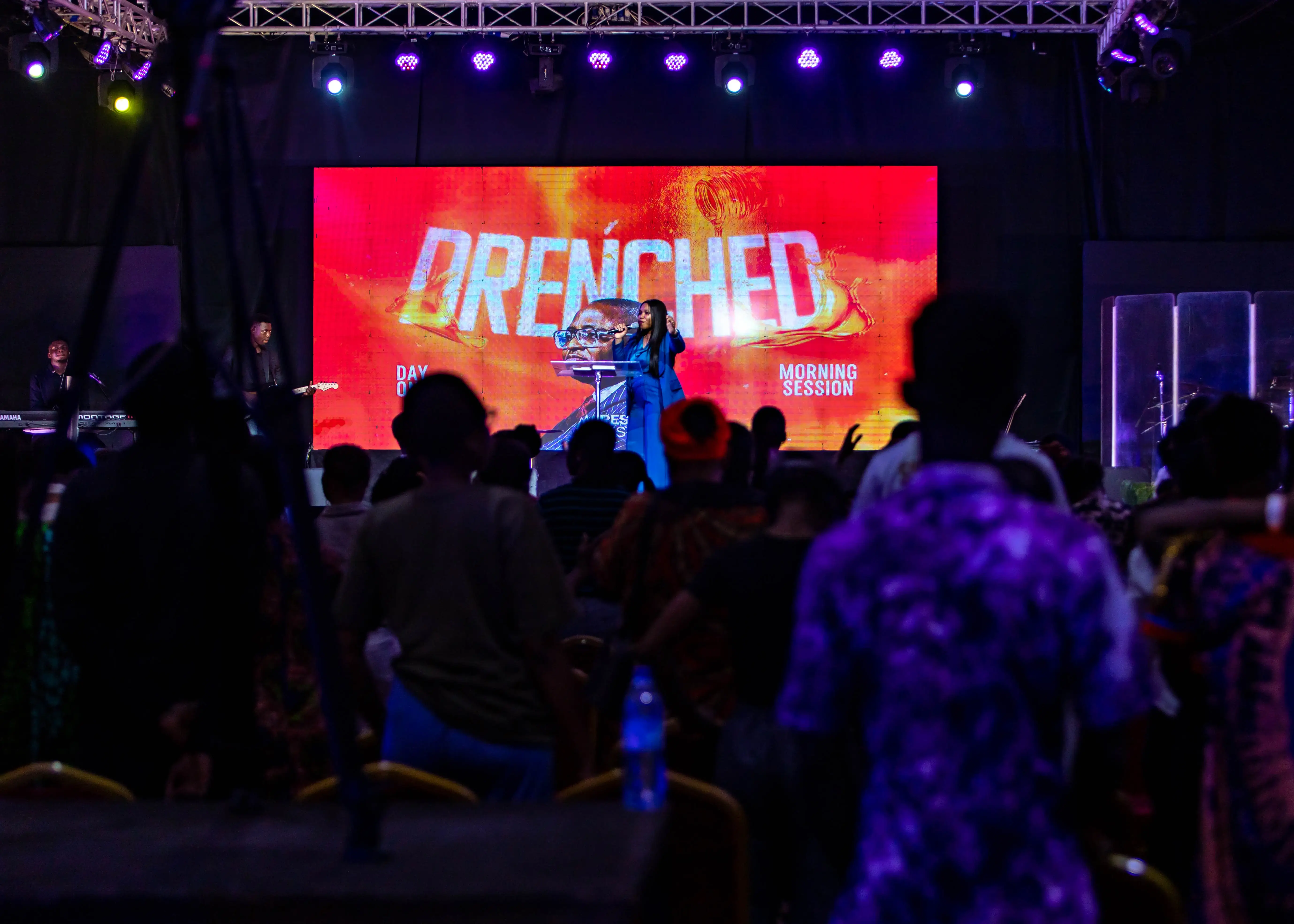 A speaker in a blue suit addressing an audience in front of a large red screen displaying the word 'DRENCHED' and text 'Day One Morning Session'.