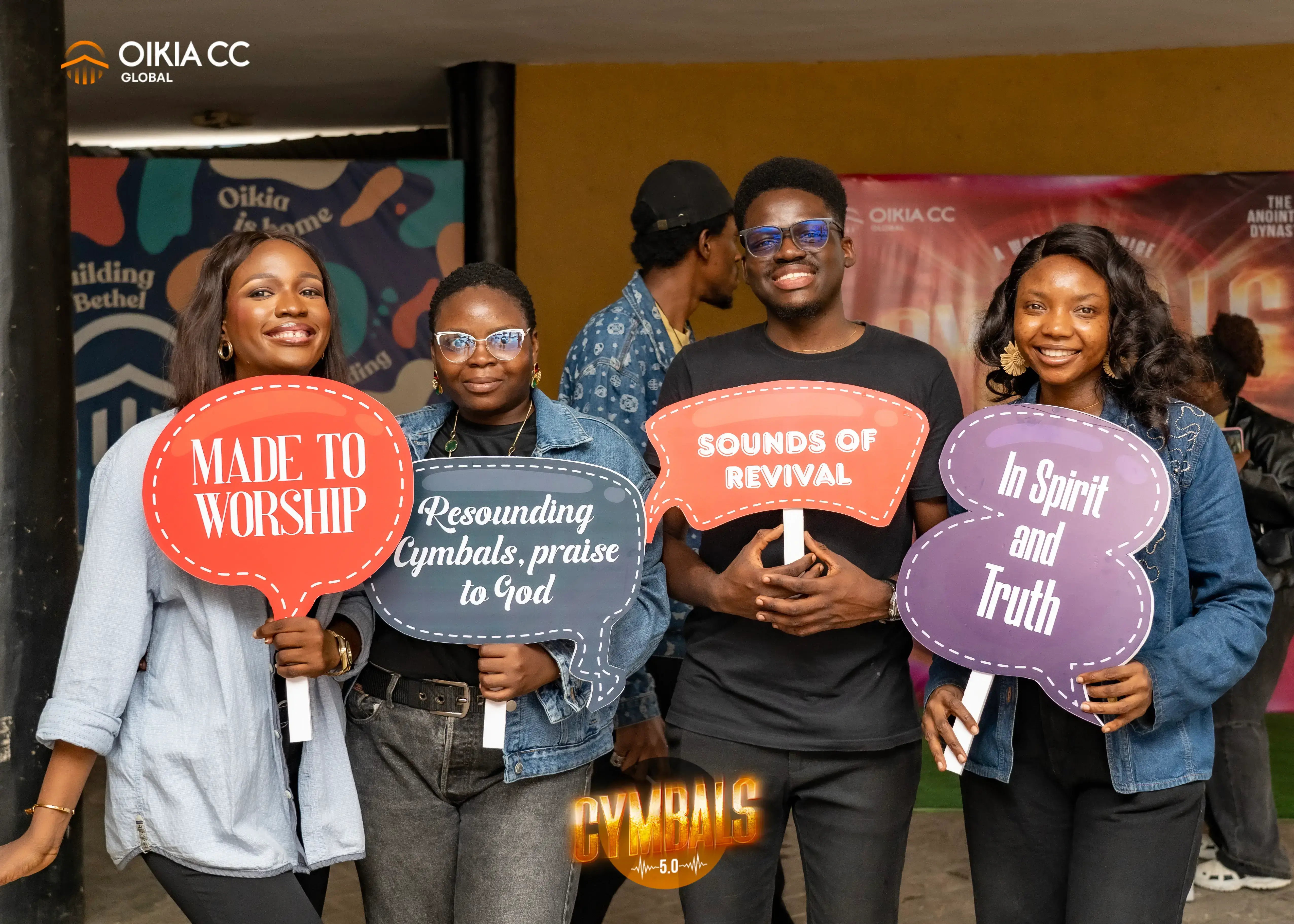Four smiling young adults holding colorful speech bubble signs with phrases about worship and praise at an Oikia CC Global event.