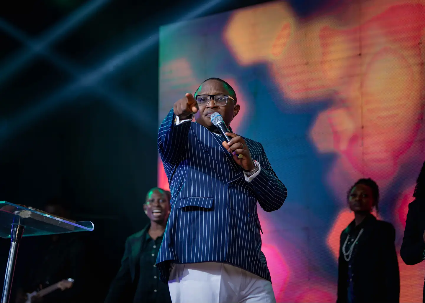 Man in a pinstripe suit speaking passionately into a microphone on stage with colorful background lights.