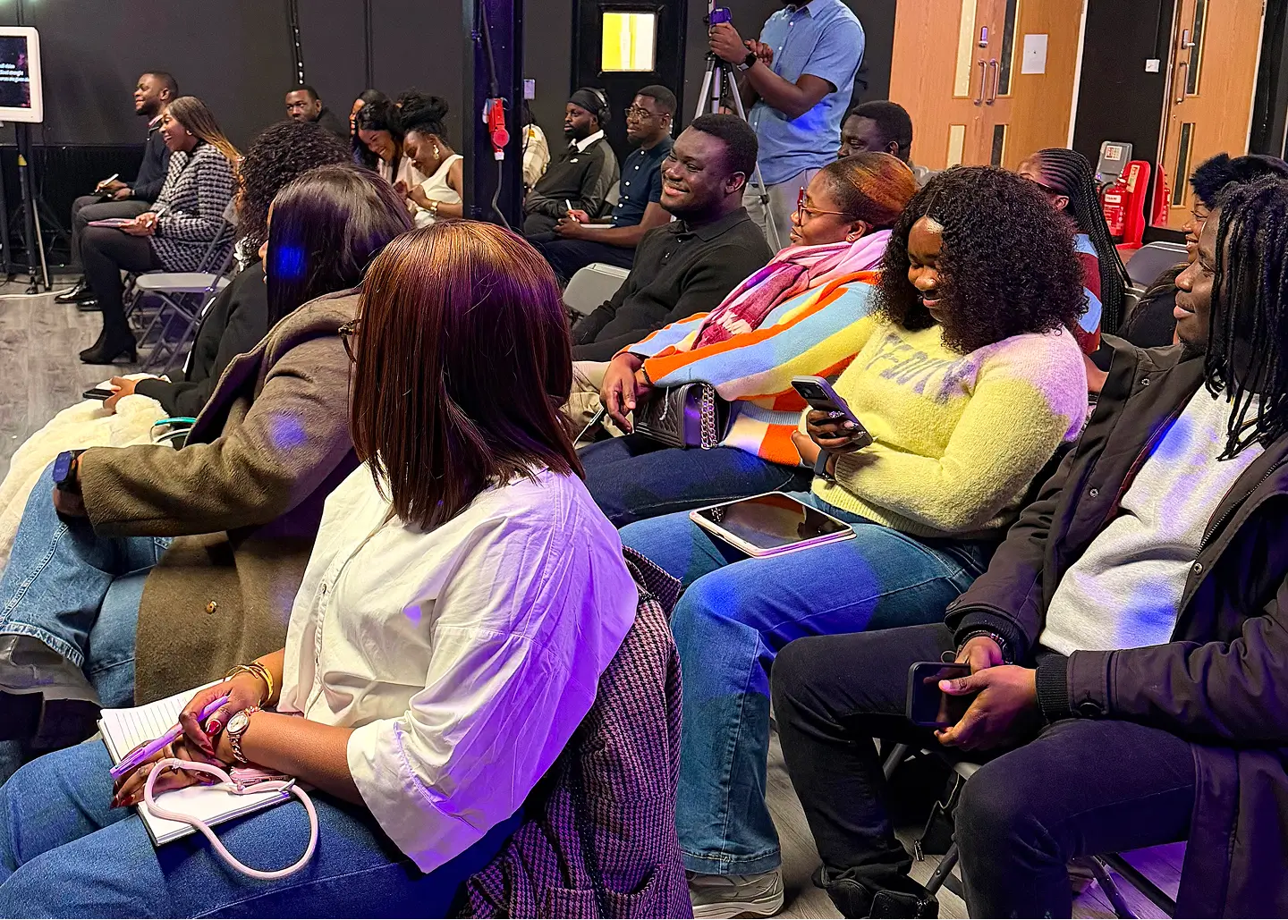 Group of people seated in rows indoors, attentively engaged, some holding phones and notebooks.