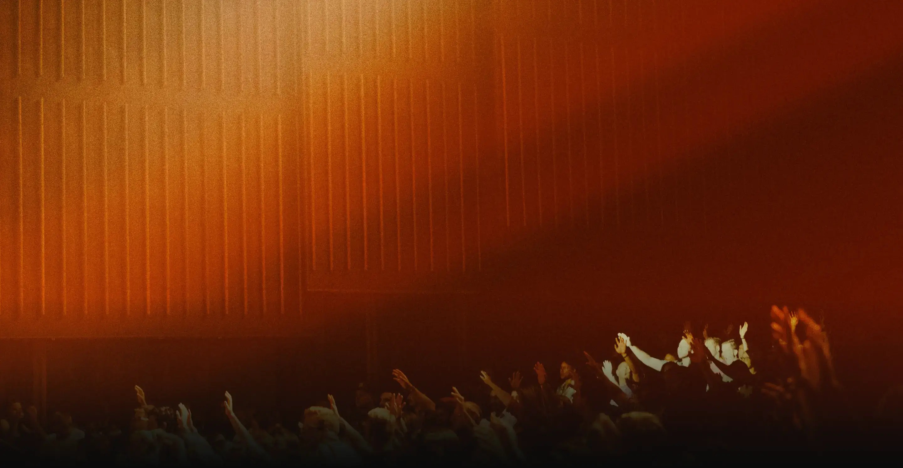 Crowd with raised hands illuminated by warm orange stage lighting at a concert or event.