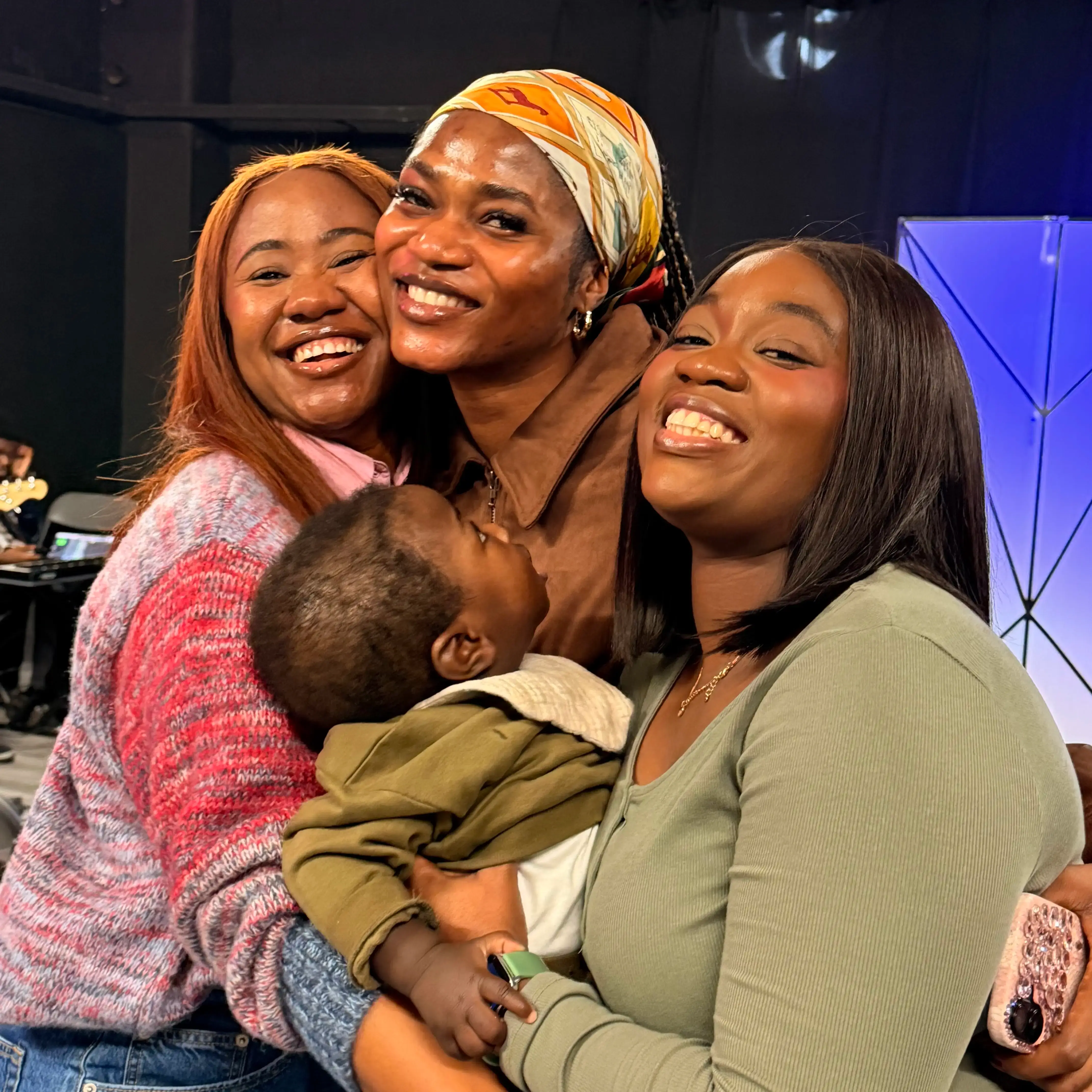 Three smiling women hugging and holding a baby in an indoor setting with a blue background.