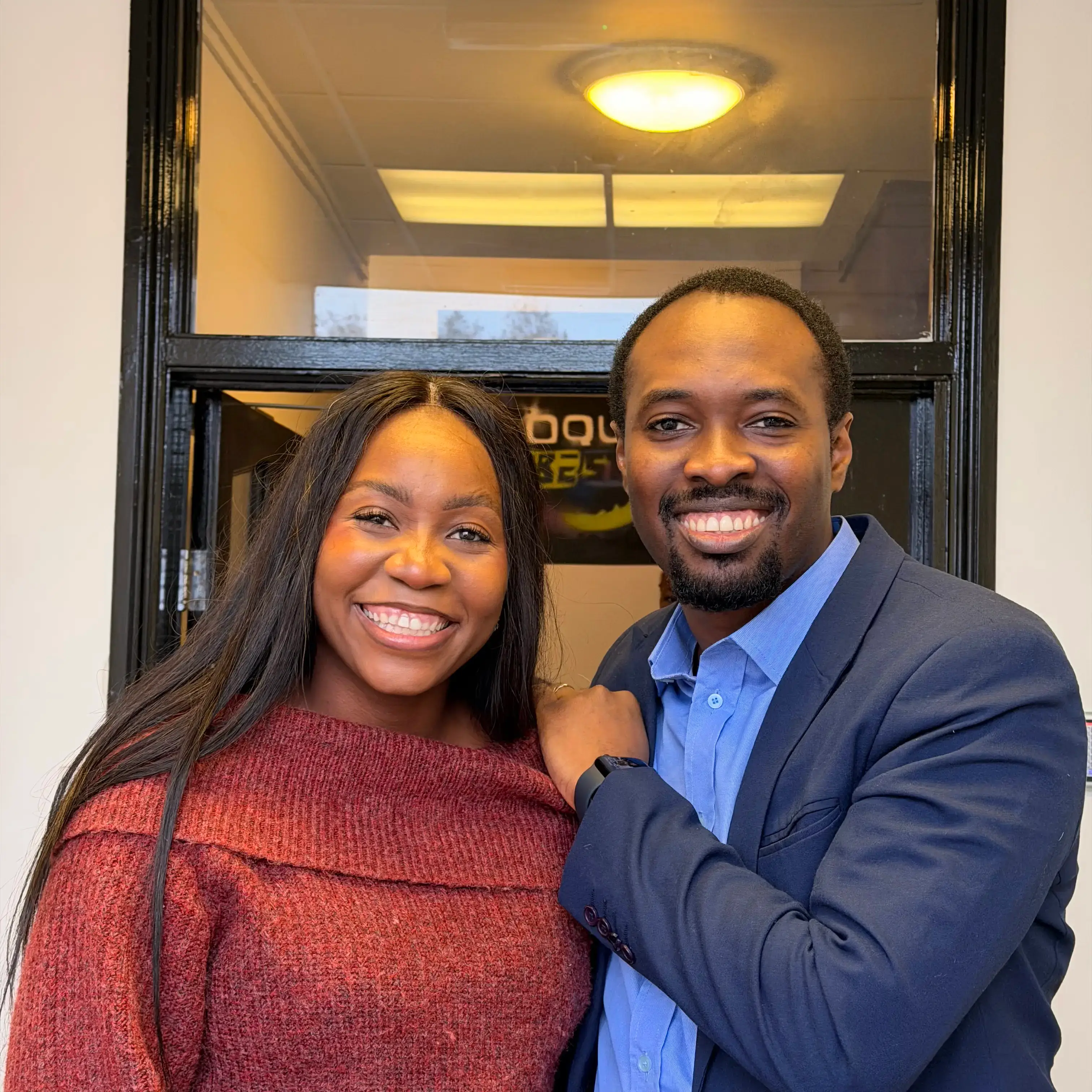 Smiling man in a blue blazer and woman in a red sweater standing closely together indoors.