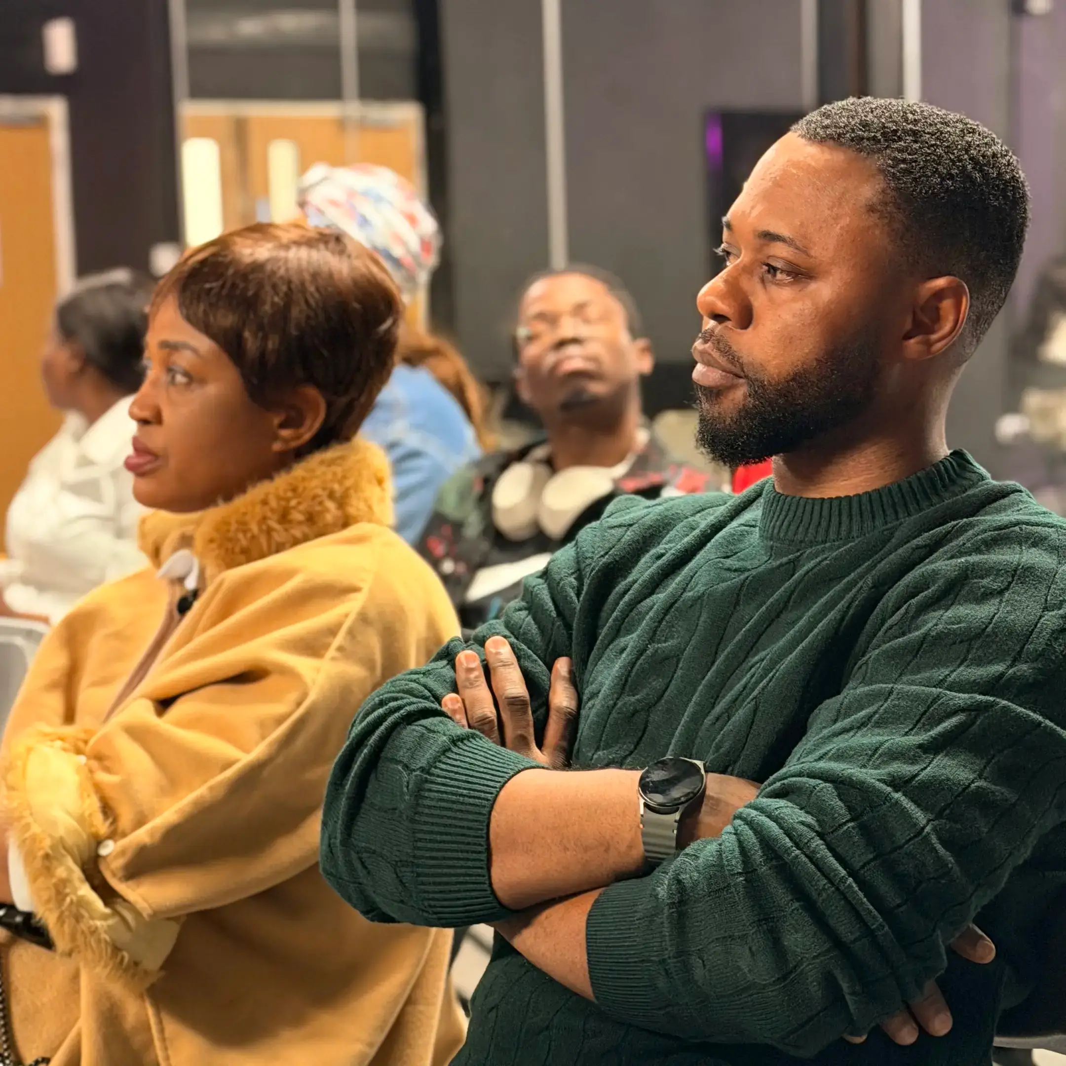 Group of adults sitting indoors attentively listening, with focus on a man in a green sweater and a woman in a tan fur-collared coat.