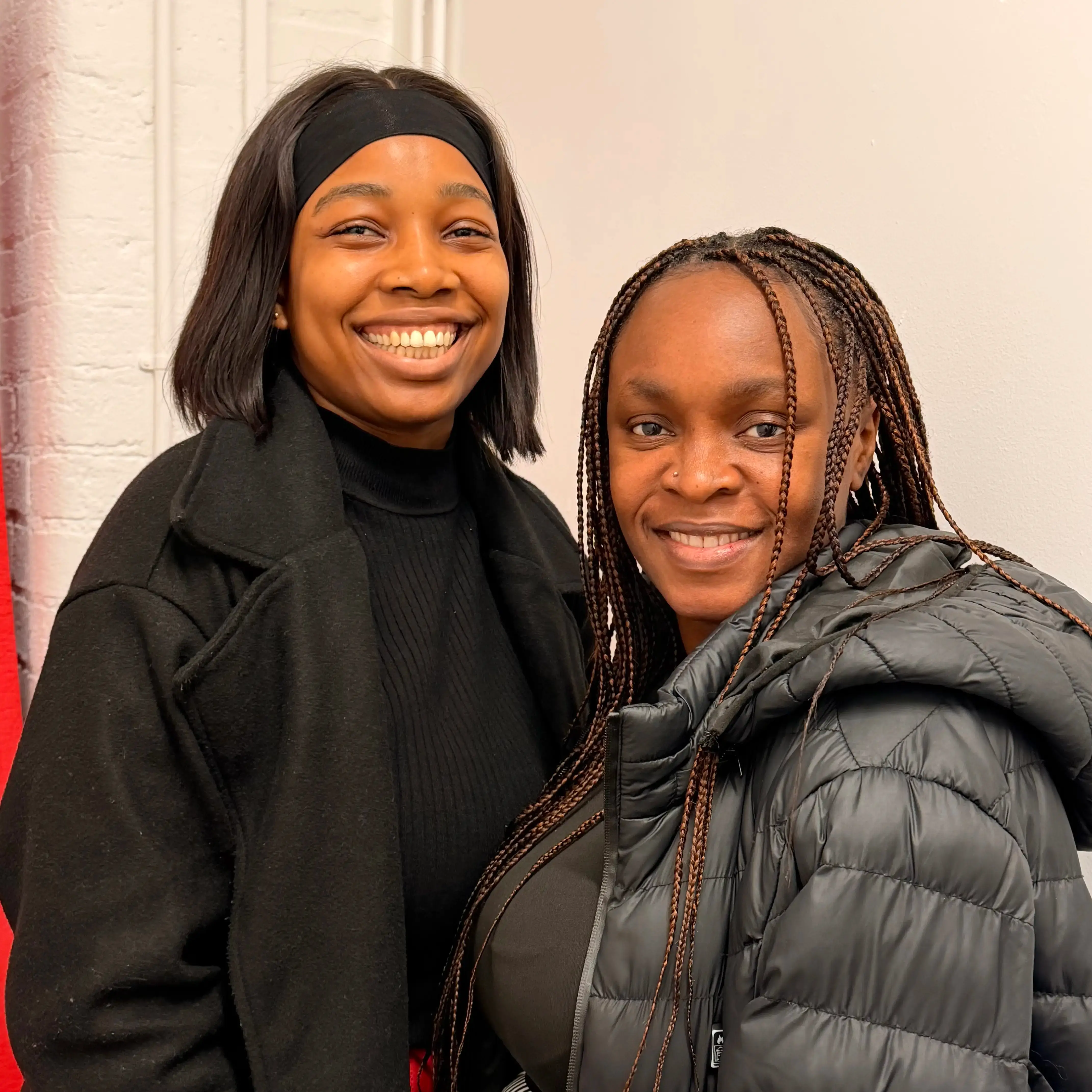 Two smiling women standing close together, one wearing a black headband and coat, the other with braided hair and a gray puffer jacket.