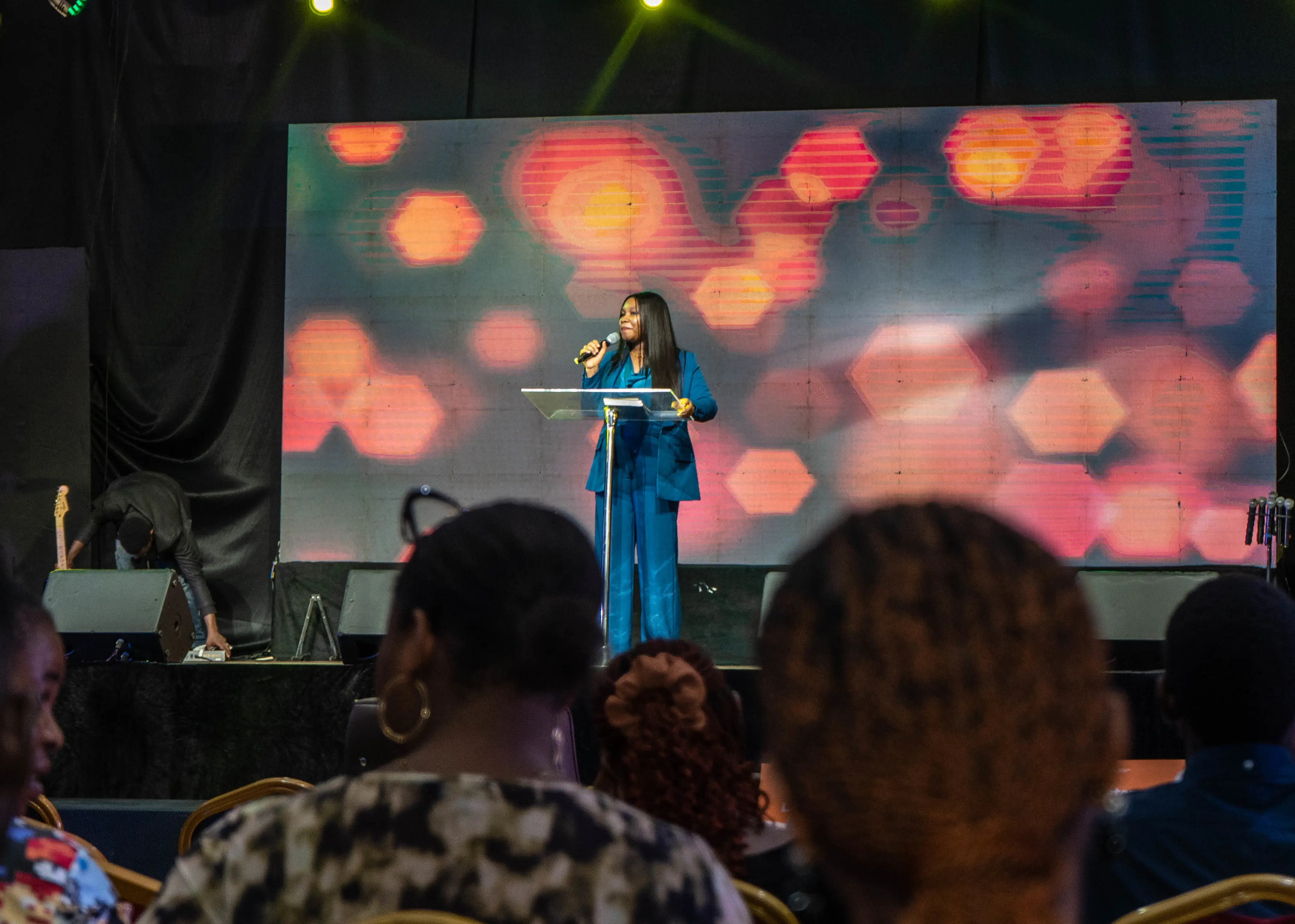 Woman in blue suit speaking into a microphone on stage with a colorful blurred background and audience in the foreground.