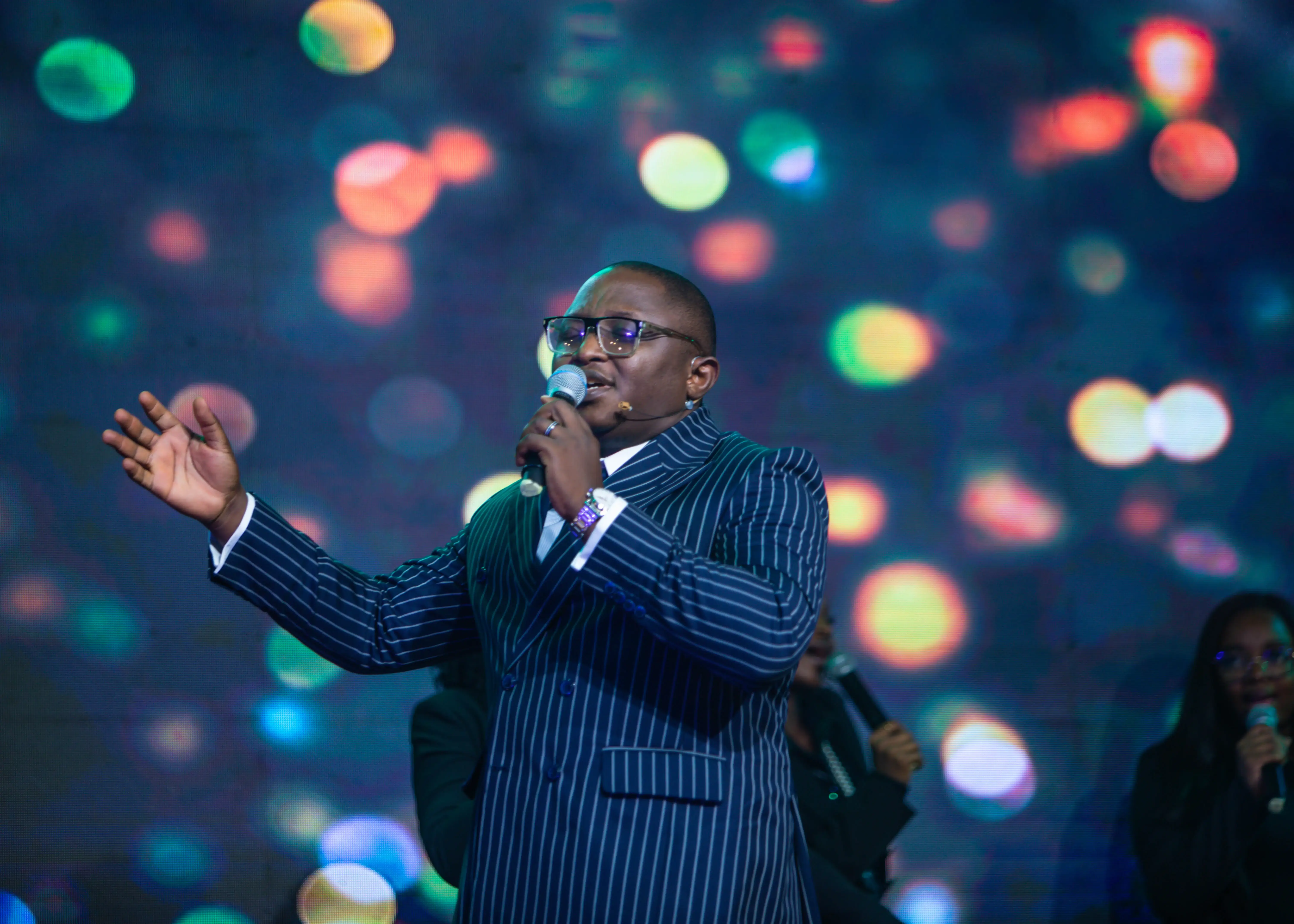 Man in a pinstripe suit singing into a microphone with colorful blurred lights in the background.