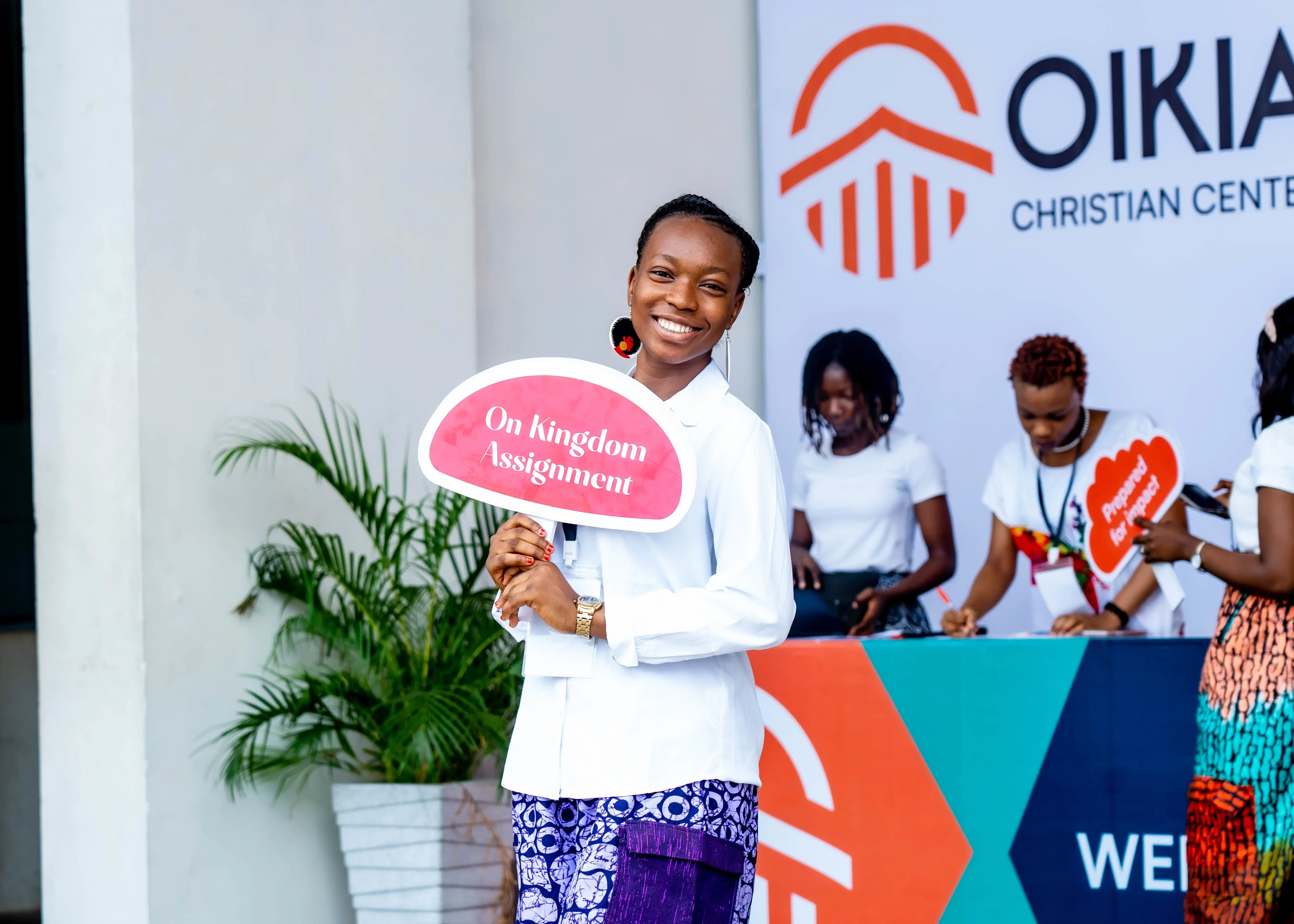 Smiling woman holding a sign that reads 'On Kingdom Assignment' with a Christian center event backdrop.