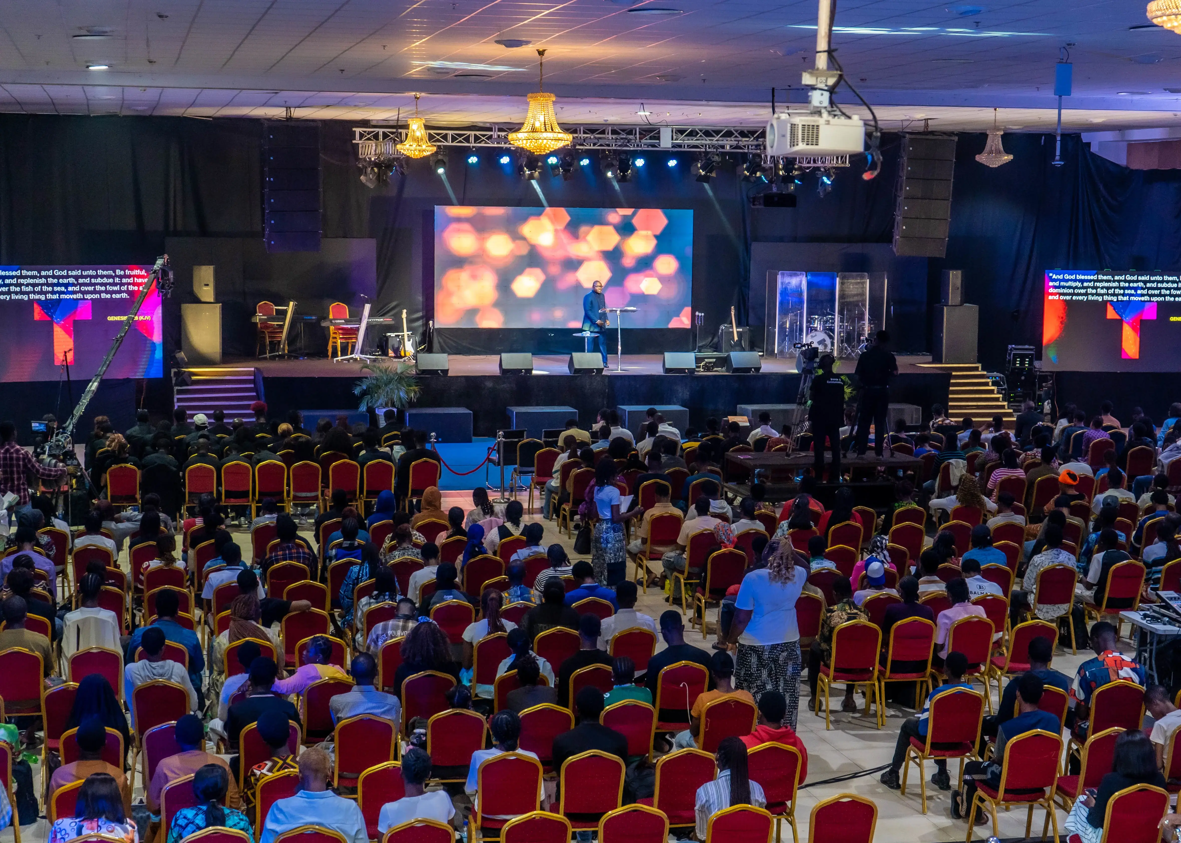 Large audience seated in rows facing a stage where a speaker stands in front of a large screen with colorful abstract visuals in a conference hall.