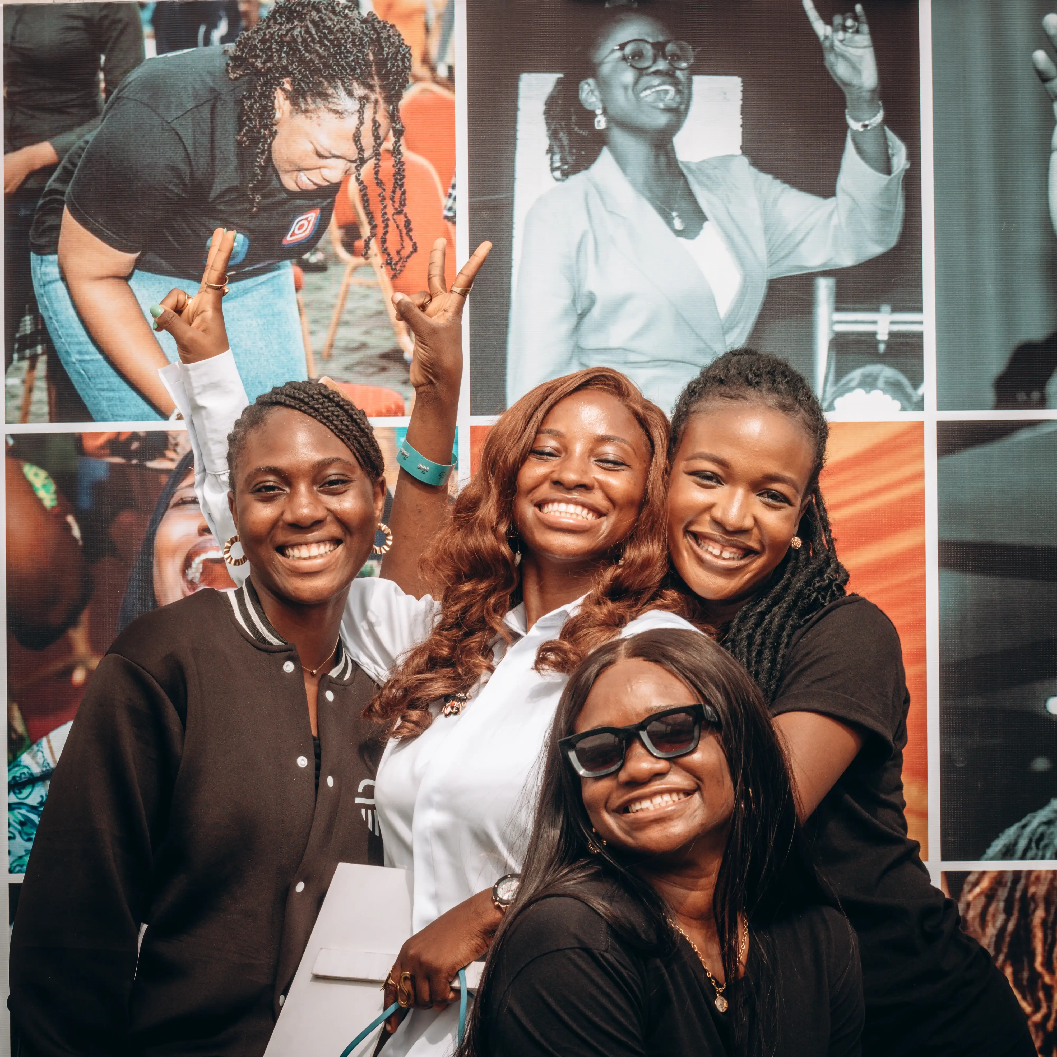 Four smiling young women posing together in front of a colorful photo collage, one wearing sunglasses.