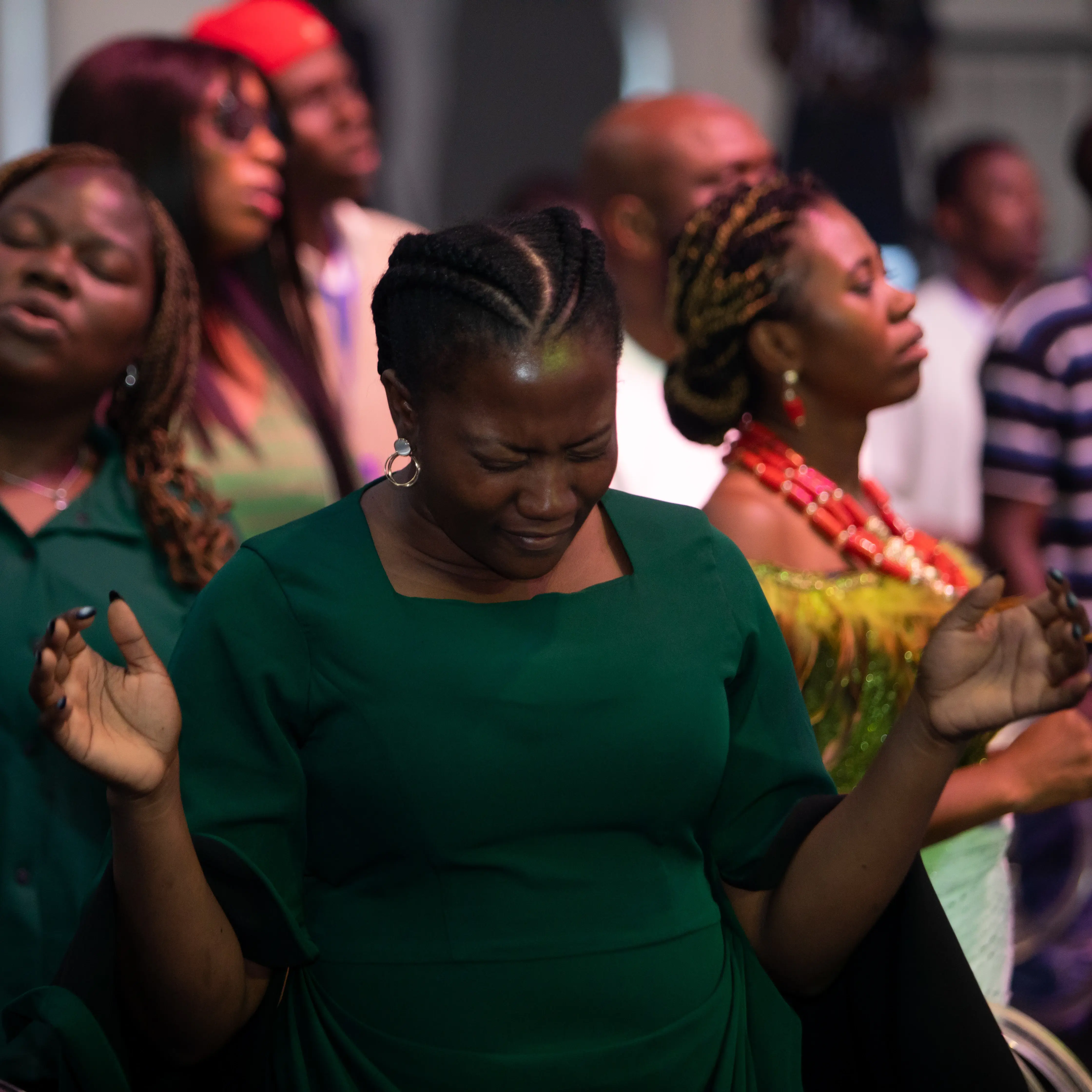 A group of people with eyes closed, appearing to be in prayer or worship, with a woman in green in the foreground raising her hands.