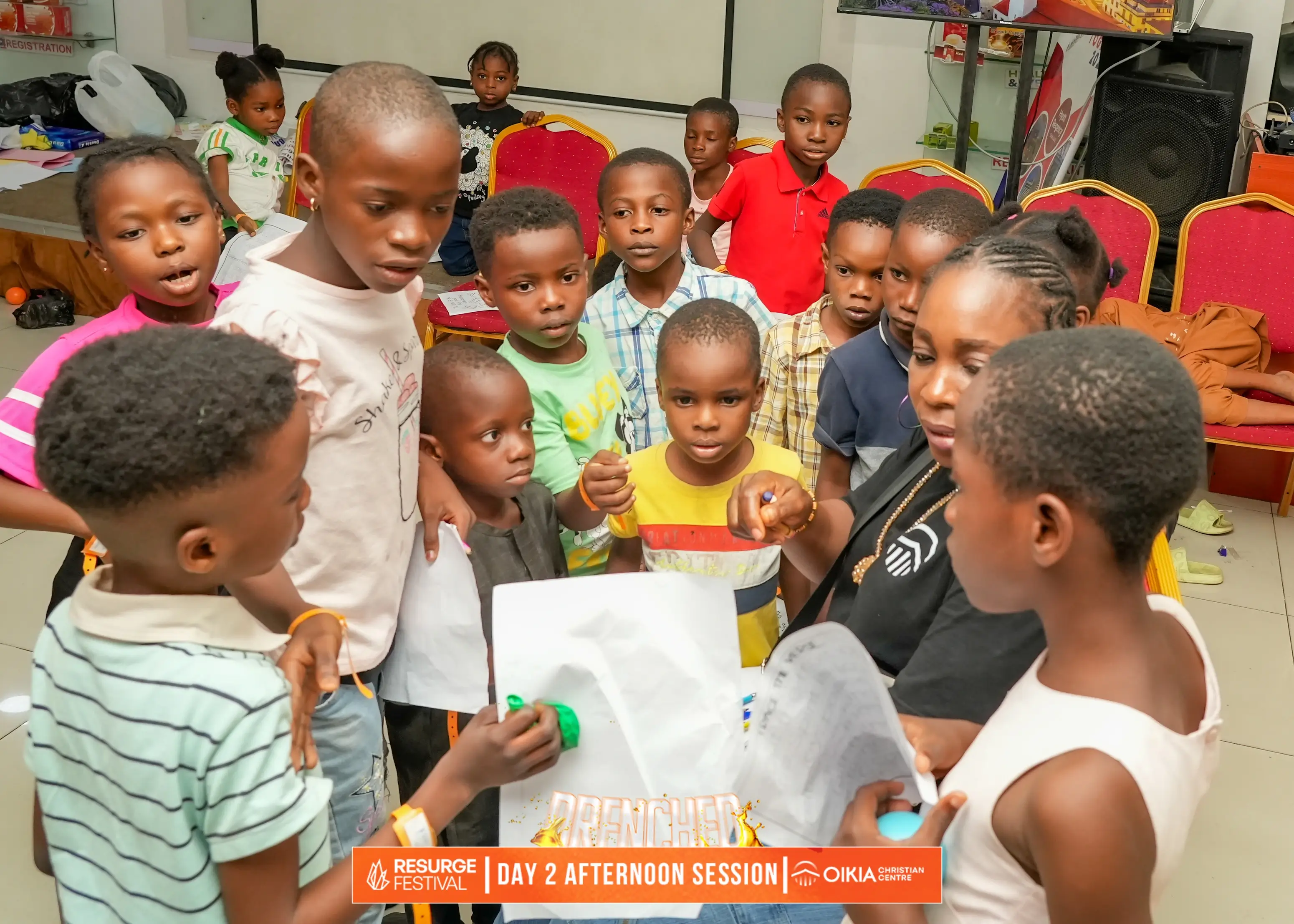 Group of children and an adult gathered closely around a document, engaged in a learning activity at Resurge Festival Day 2 Afternoon Session.