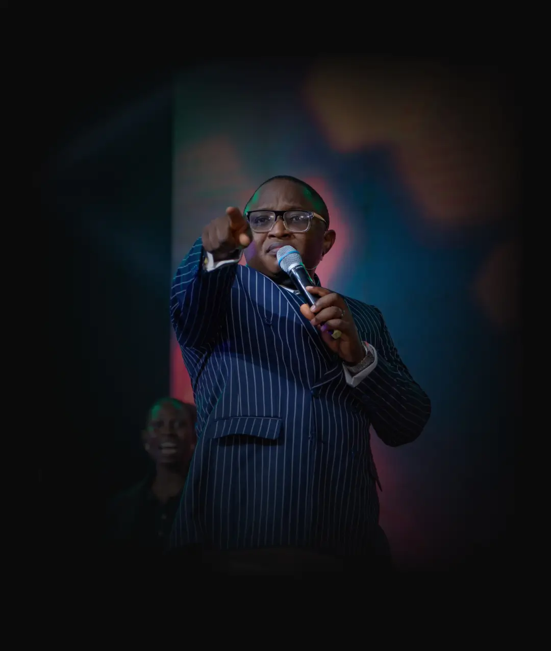 Man in pinstripe suit and glasses holding a microphone, pointing forward with a serious expression.