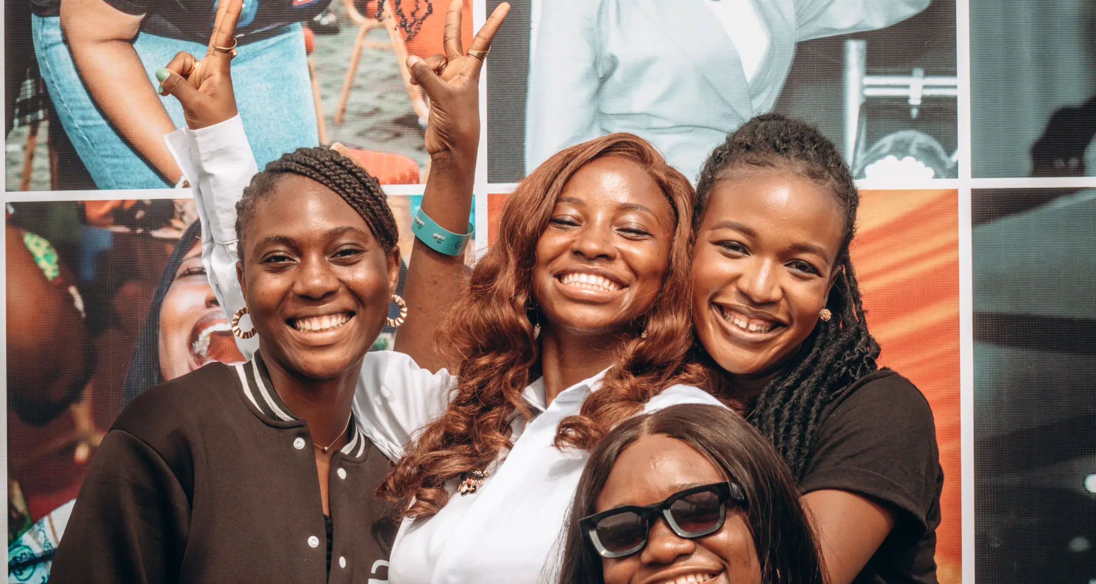 Four smiling women pose closely together, one making peace signs with both hands.