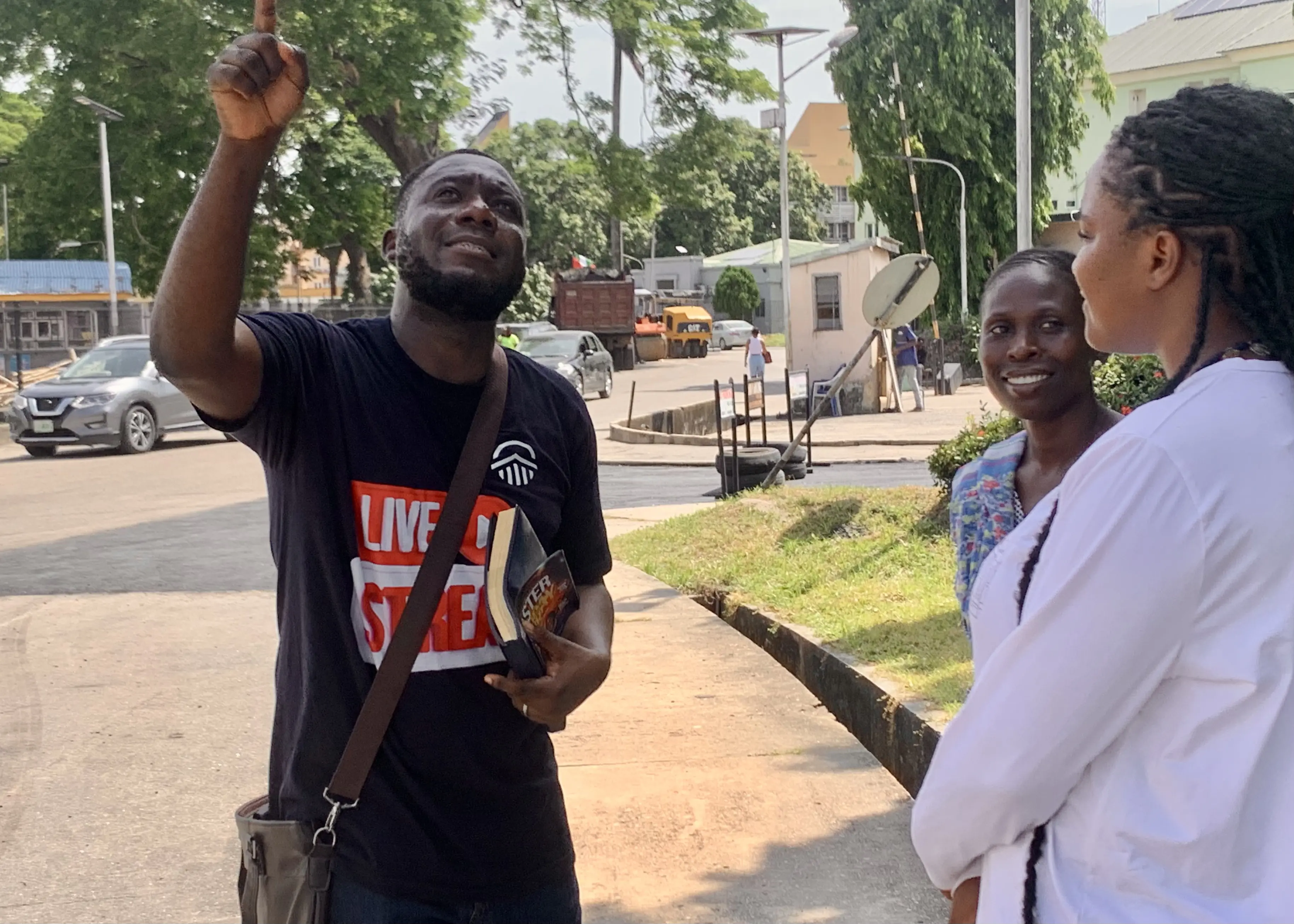 Man in Live On Street shirt holding a book and pointing upward while talking to two women outside.