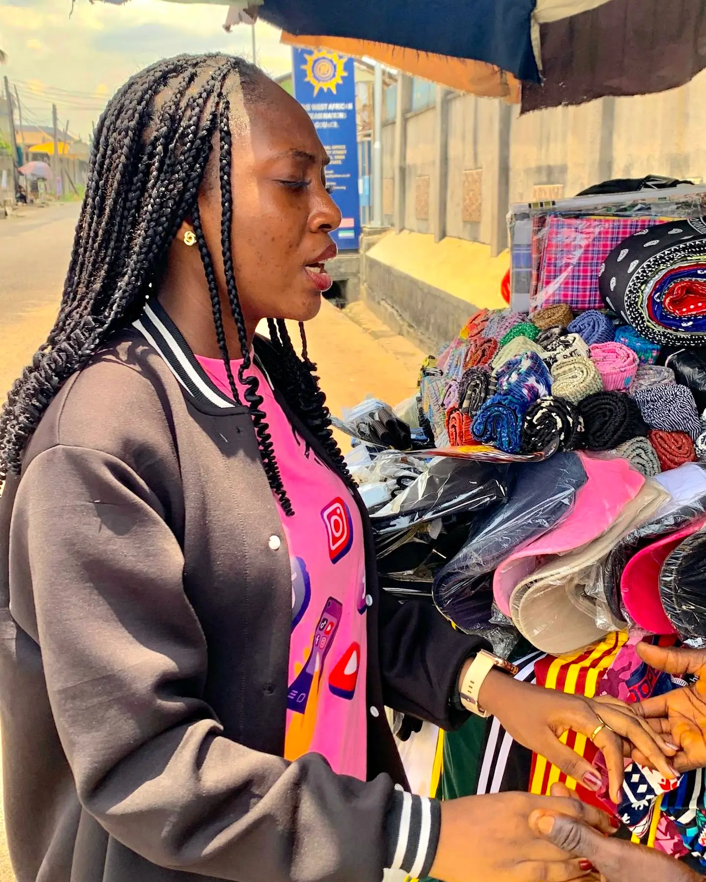 Woman with braids in a black jacket buying colorful fabric at an outdoor market stall.