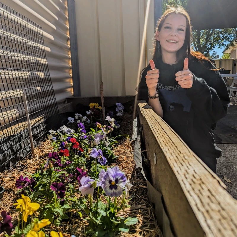 Student posing with two thumbs up next to colourful garden bed of flowers. 
