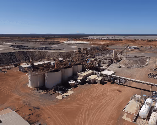 Aerial view of an industrial mining site with processing equipment and large piles of extracted materials under a clear blue sky.