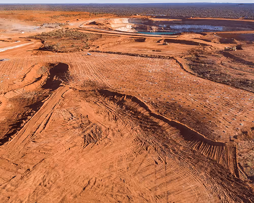 Aerial view of a large open excavation site with reddish soil and earth-moving equipment in the distance.