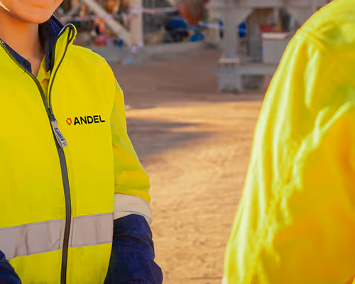 Two individuals wearing yellow high-visibility jackets with one jacket displaying the ANDEL logo.