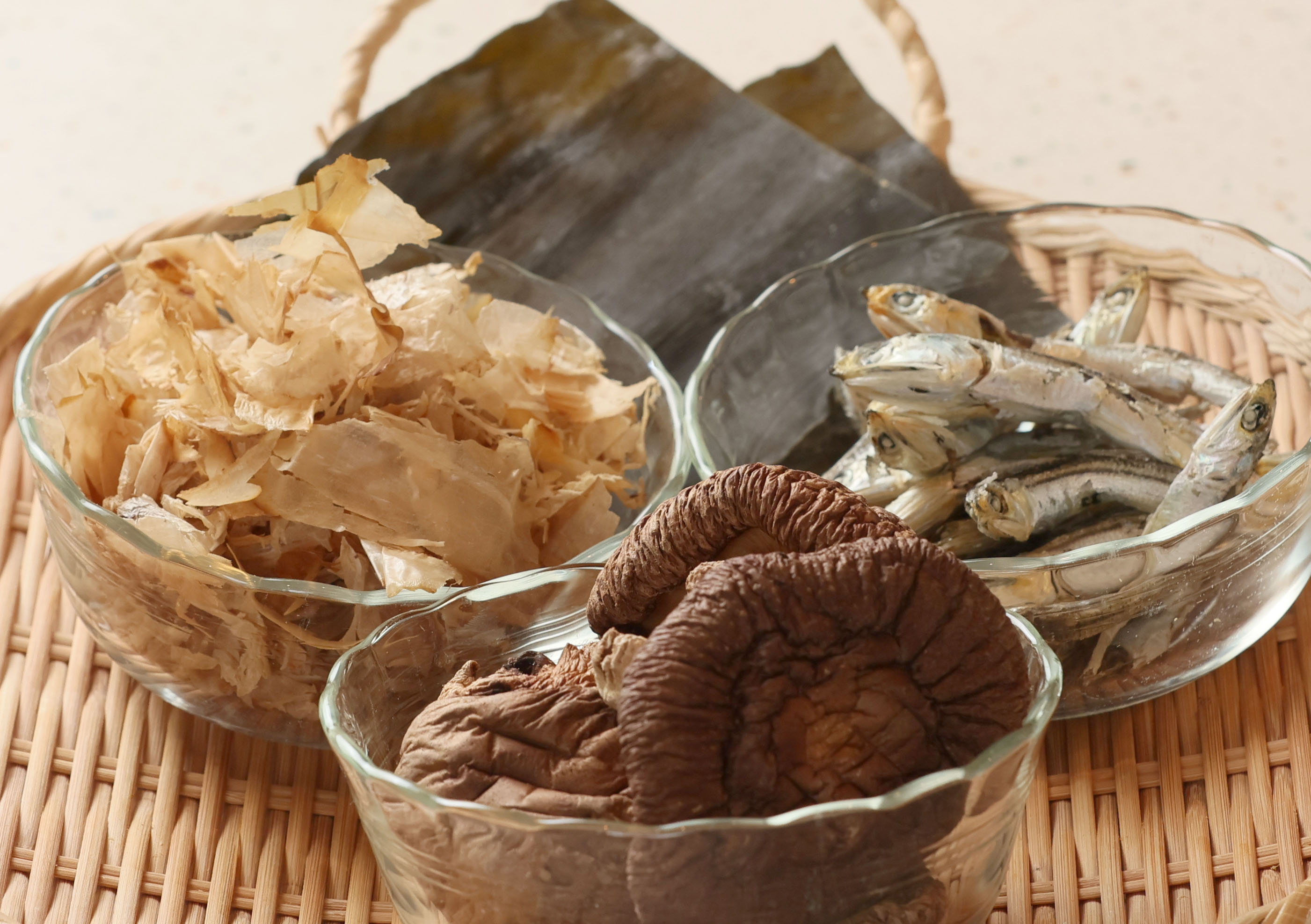 Four Japanese dashi ingredients arranged in glass bowls — katsuobushi bonito flakes, dried kombu seaweed, niboshi dried anchovies, and dried shiitake mushrooms — displayed together in a wicker basket.