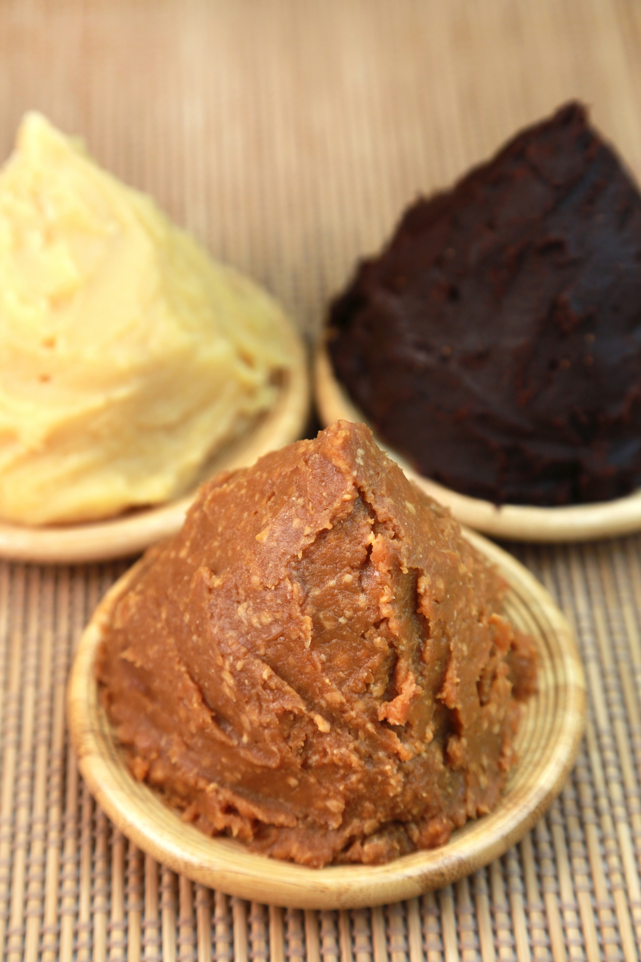 Three types of Japanese miso paste — pale yellow shiro miso, reddish-brown medium miso, and deep dark aka miso — each mounded on a small bamboo plate, arranged on a woven mat.