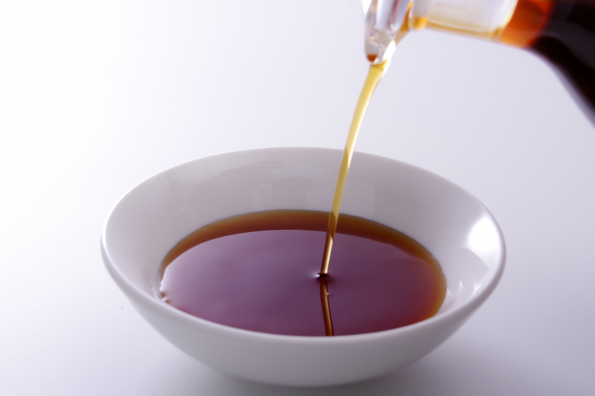 Dark Japanese soy sauce being poured in a steady stream into a round white ceramic dish, photographed against a clean white background.