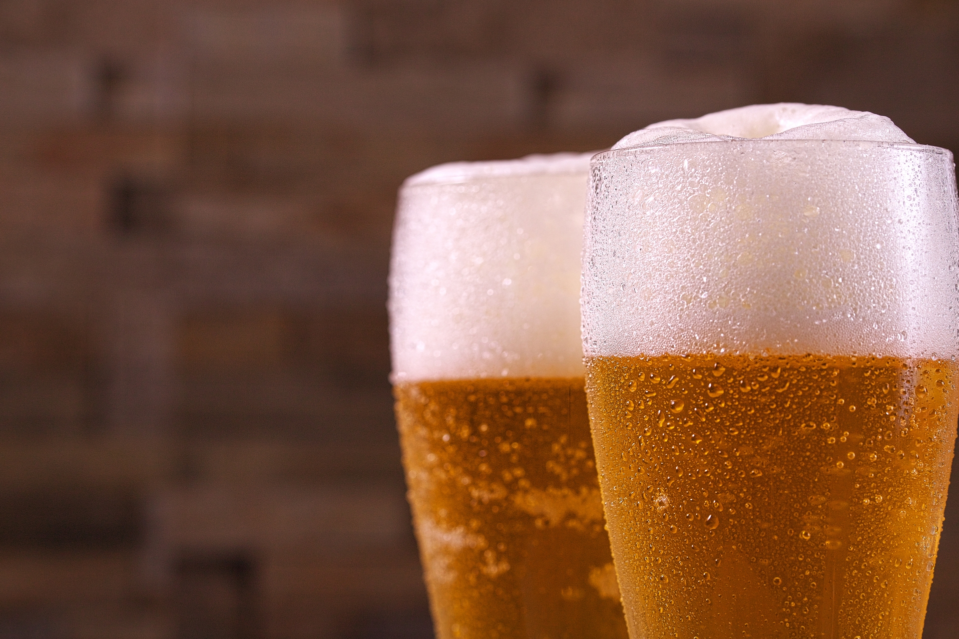 Two ice-cold glasses of golden Japanese beer with thick white foam heads, beaded with condensation, photographed up close against a warm brick wall background.