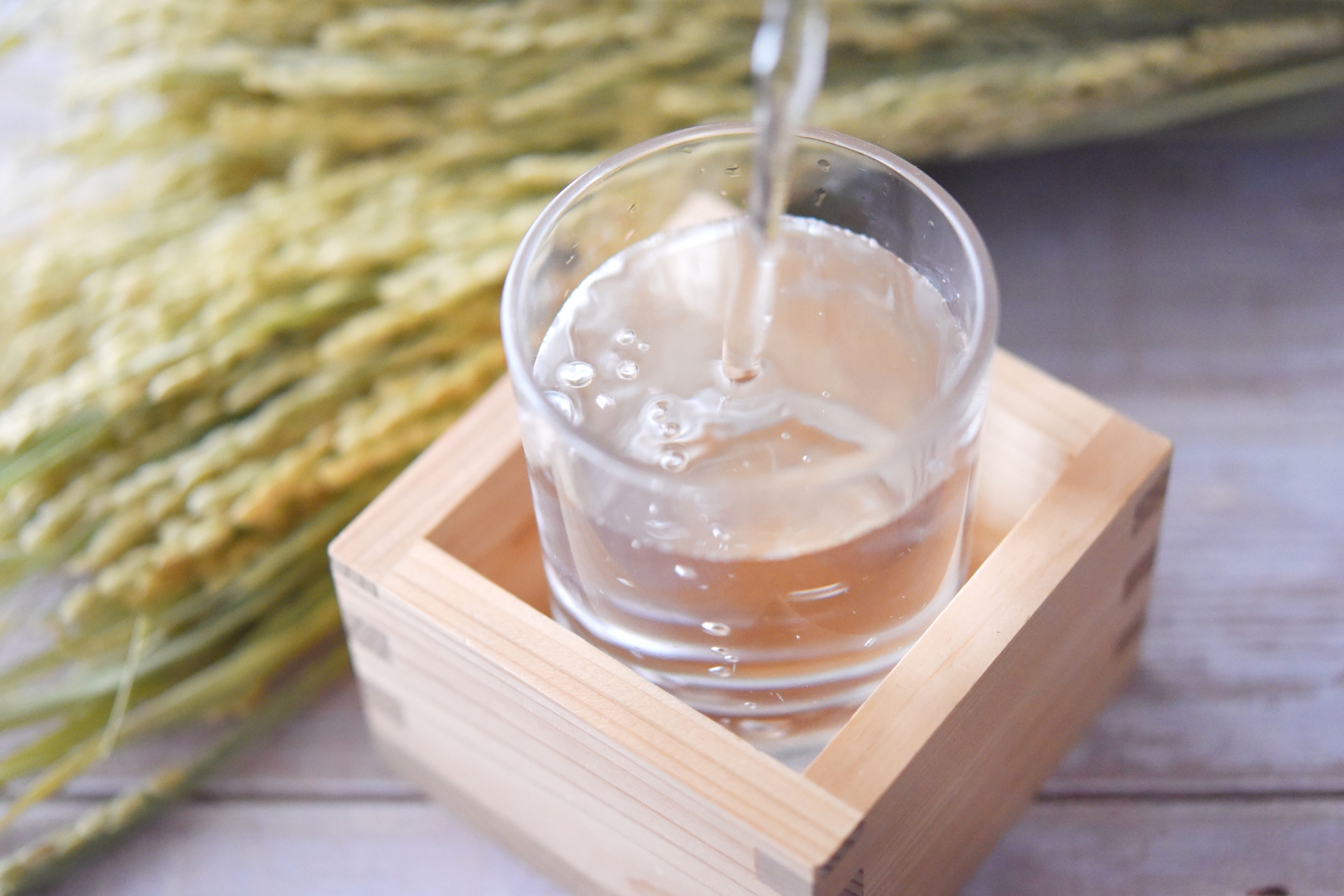 Golden sake being poured into a glass nestled inside a wooden masu, overflowing in the mokkiri style, with soft-focus grain stalks in the background.
