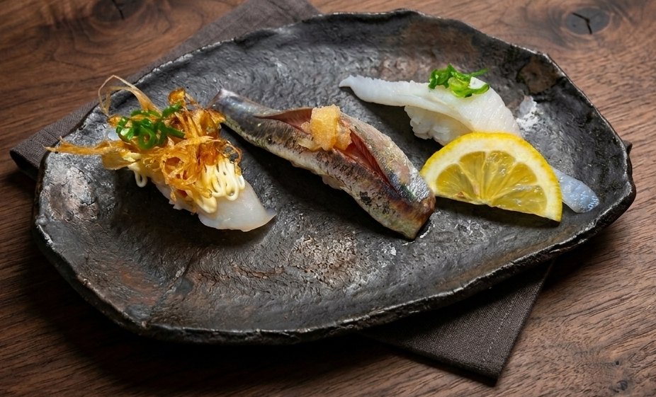 Three nigiri on a dark ceramic plate — squid with fried leek and mayo, sardine, and engawa with lemon and scallion.