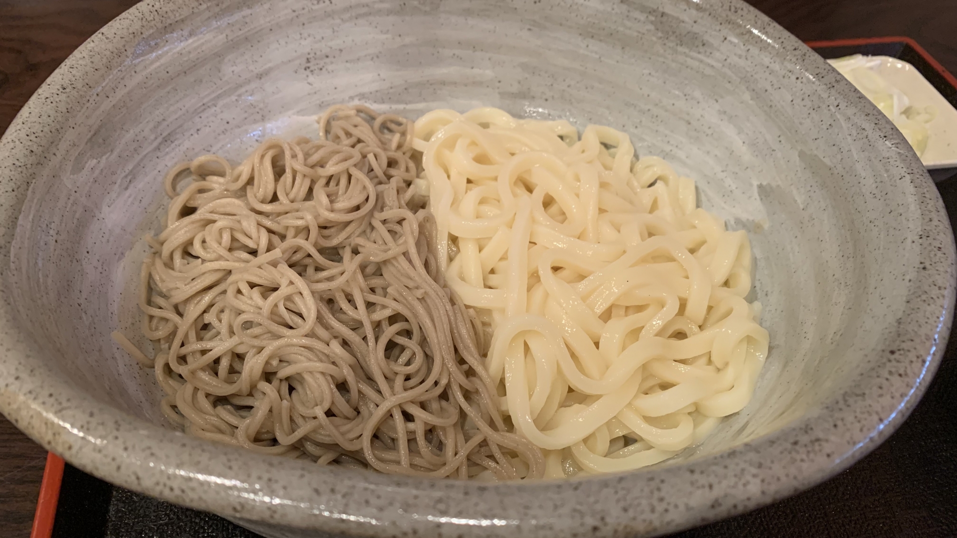 Gray buckwheat soba and white udon noodles served side by side in a single speckled ceramic bowl, on a dark wooden table.