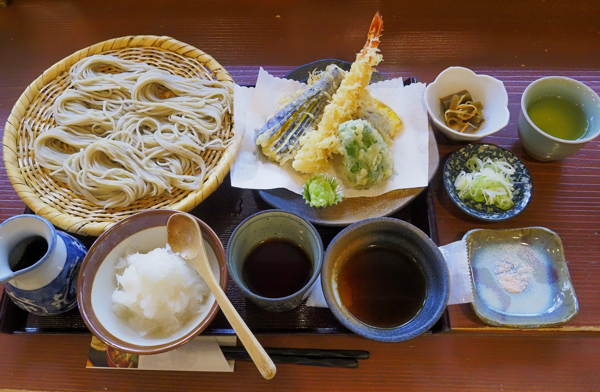 Cold soba on a bamboo zaru, shrimp and vegetable tempura, dipping sauces, grated daikon, and green tea on a dark tray.