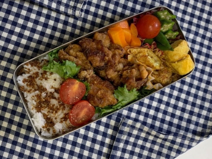 A Japanese bento with karaage fried chicken, tamagoyaki, rice with furikake, cherry tomatoes, and vegetables on a checkered cloth.