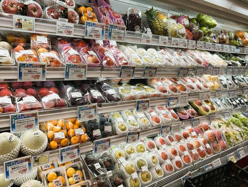 A Japanese supermarket fruit section packed with apples, peaches, mikan, melon, grapes, plums, and tropical fruits, all wrapped and labeled.
