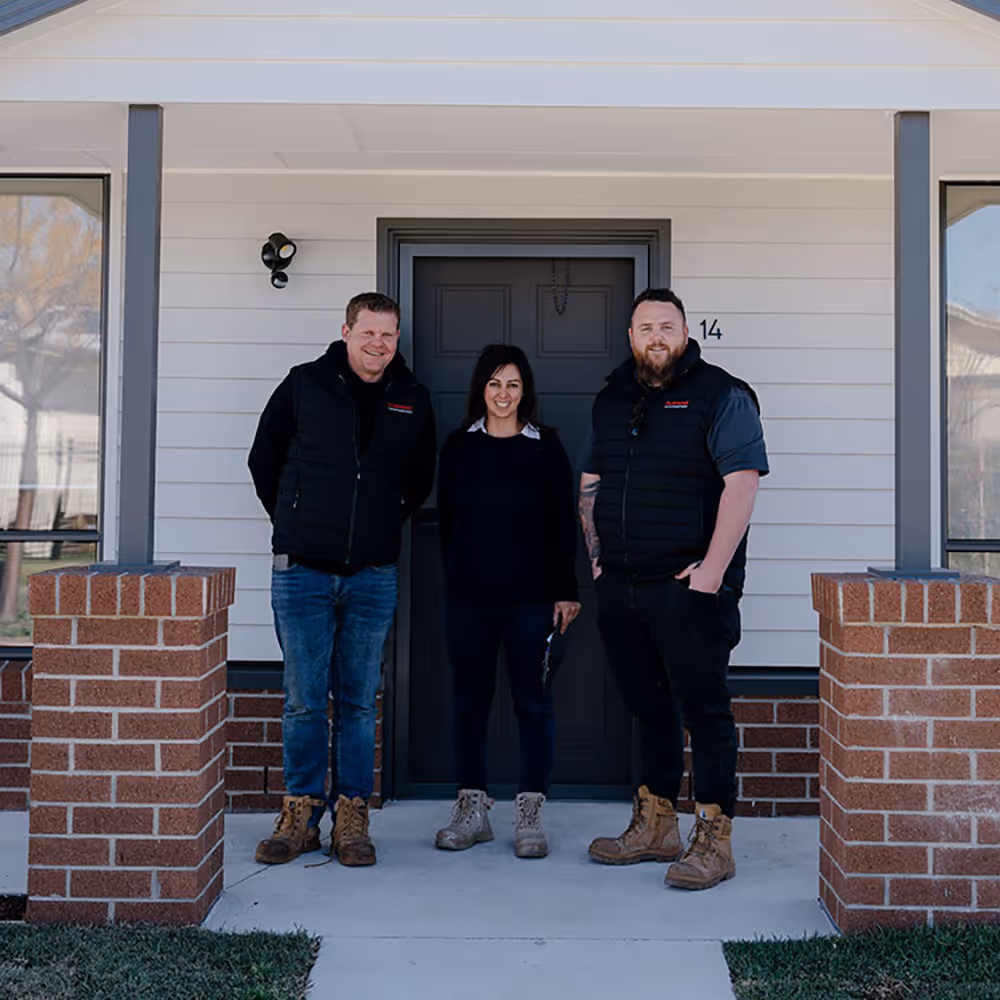 Three people standing and smiling in front of a modern house entrance with brick pillars and a gray door marked number 14.