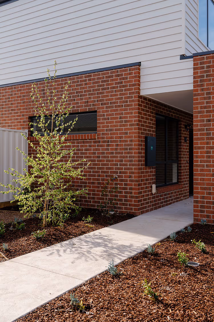 Modern house exterior showing red brick lower facade with white siding above, a narrow window, young tree, and concrete walkway.