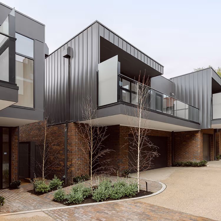 Modern townhouse with dark metal cladding upper level and brick lower level, featuring glass balcony and small landscaped area with young trees.