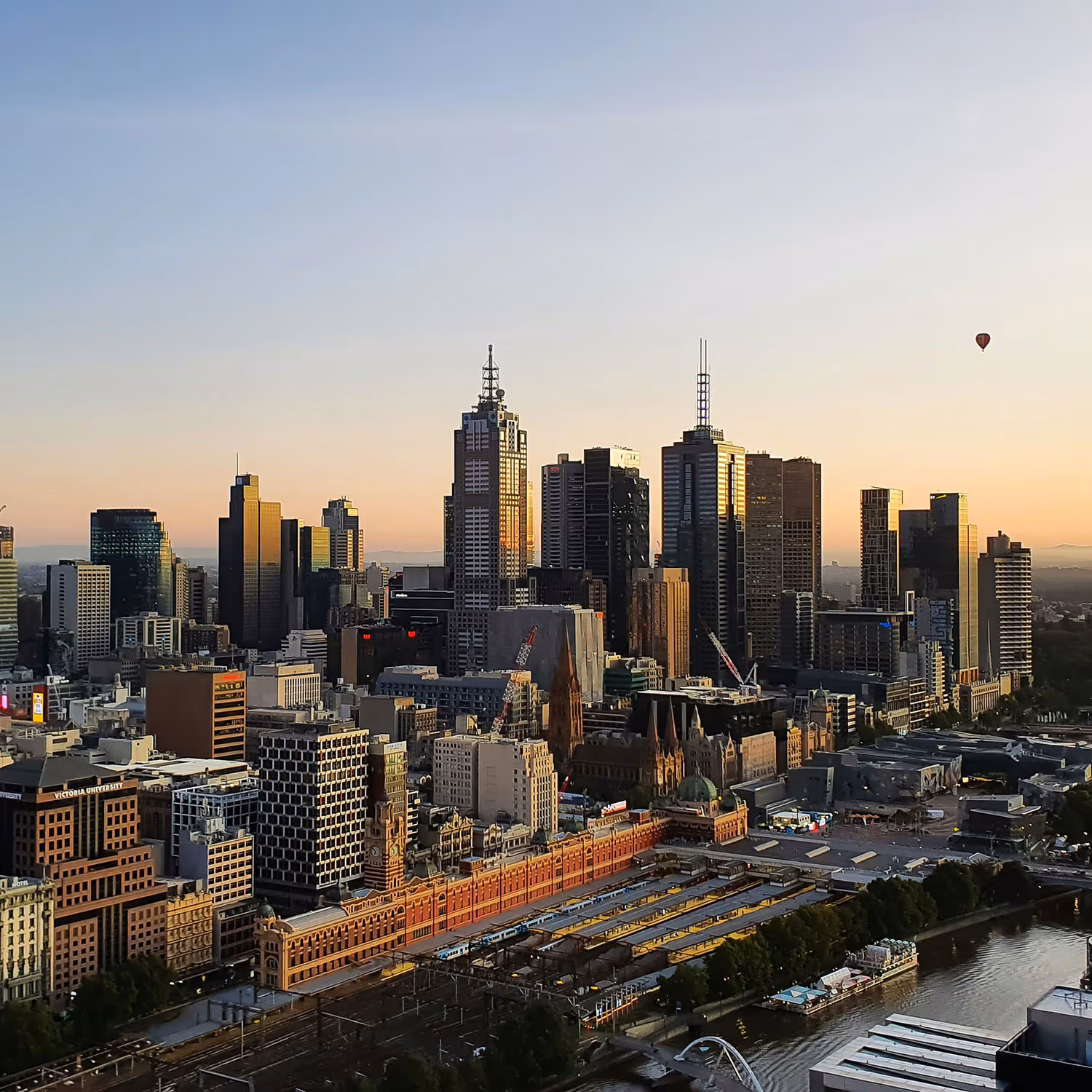 Melbourne city skyline at sunset with tall buildings, Flinders Street Station in the foreground, and a hot air balloon in the sky.