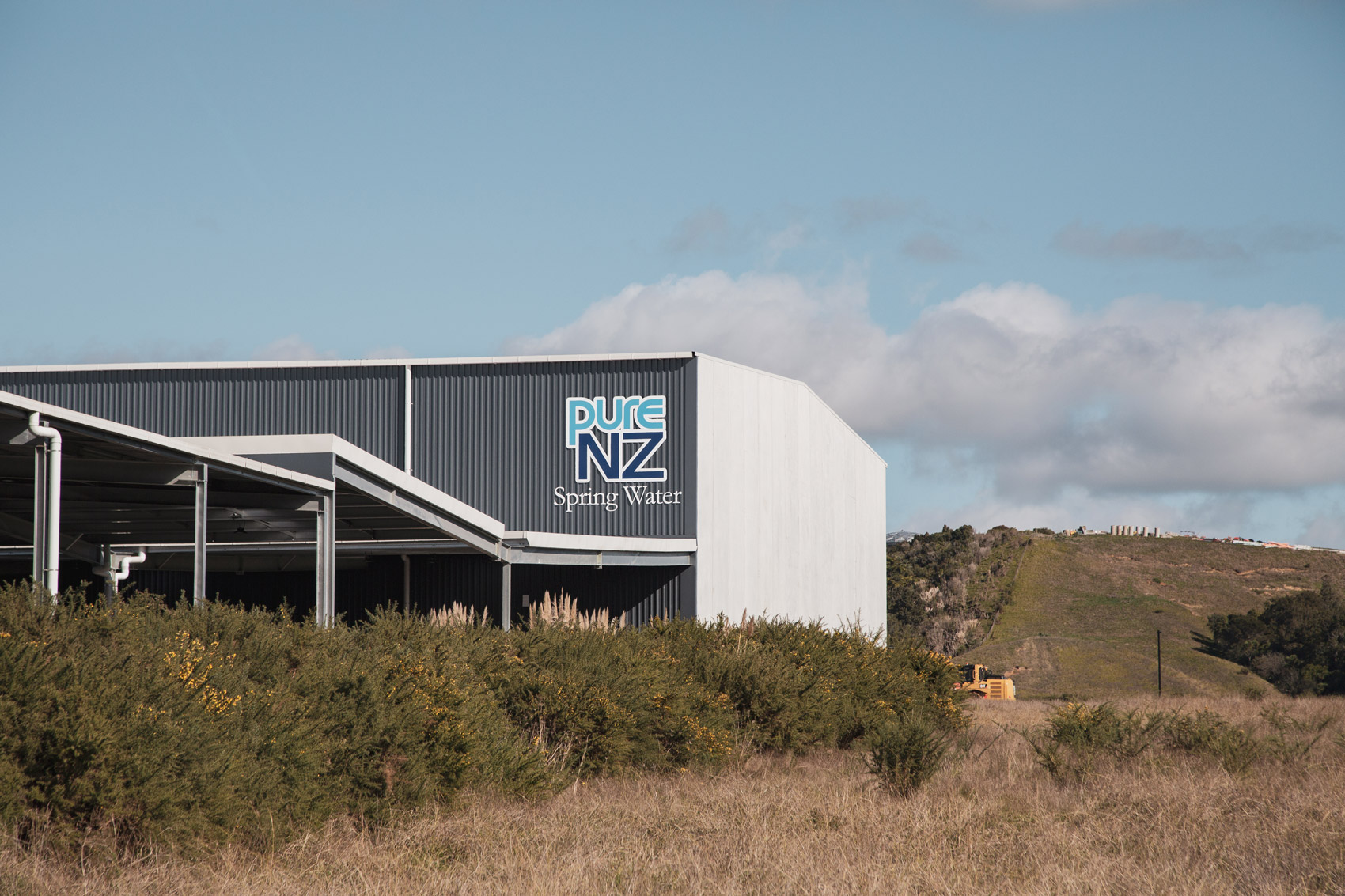 Industrial building with Pure NZ Spring Water logo, surrounded by grassy and bushy landscape under a partly cloudy sky.