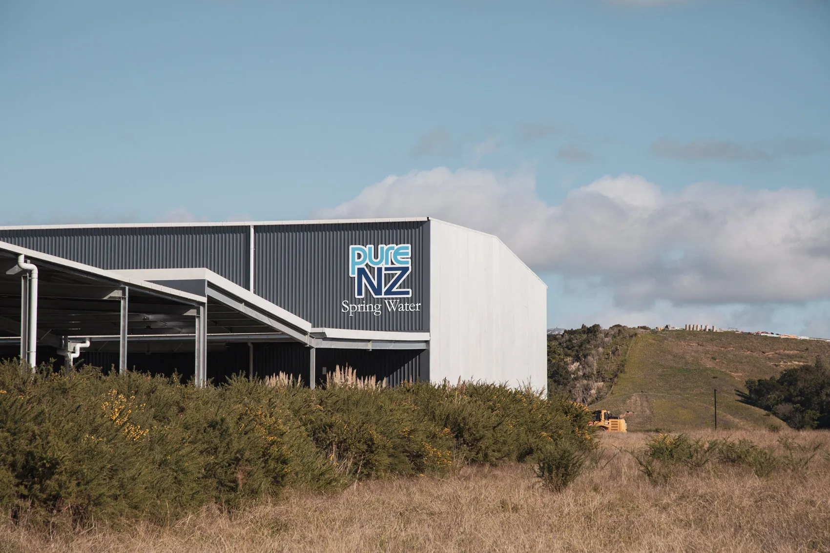 Industrial building with Pure NZ Spring Water logo, surrounded by grassy and bushy landscape under a partly cloudy sky.