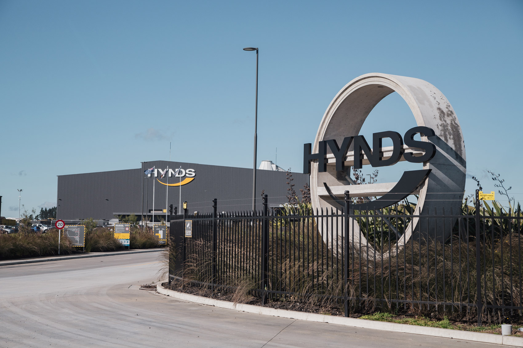 Entrance to Hynds industrial facility with circular concrete logo structure and large building under clear blue sky.