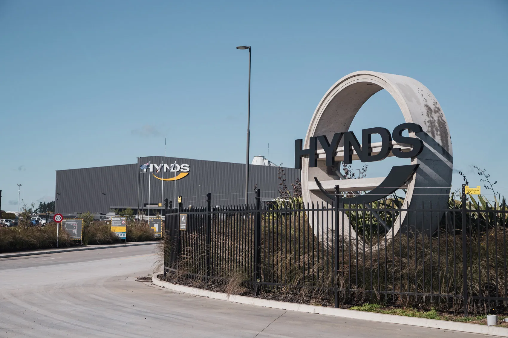 Entrance to Hynds industrial facility with circular concrete logo structure and large building under clear blue sky.
