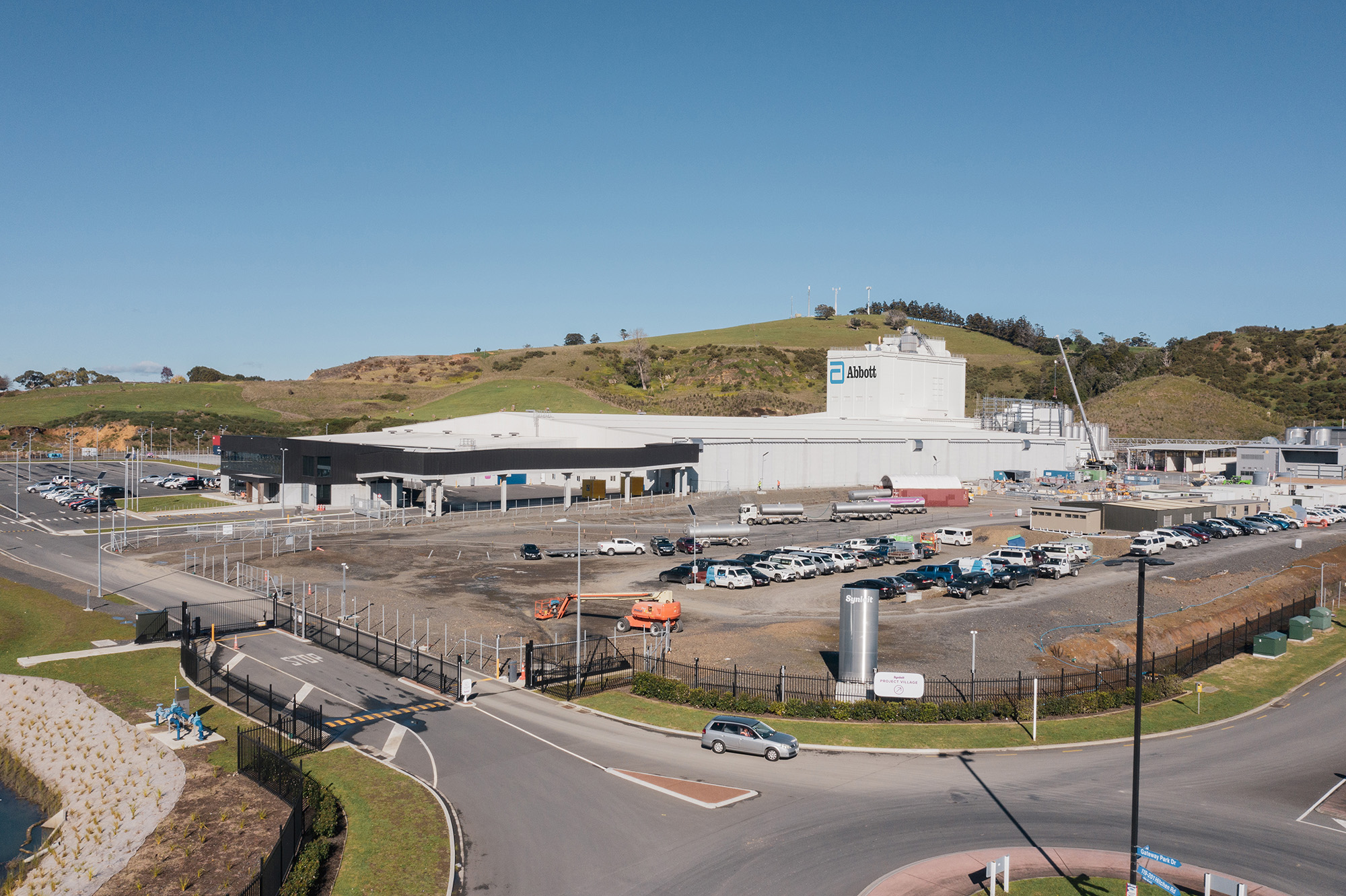 Wide aerial view of the Abbott Laboratories NZ facility with parked cars, fencing, and green hills in the background under a clear blue sky.