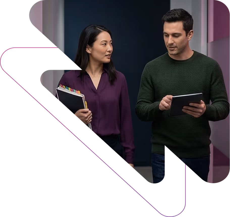 A woman holding file folders talks to a man using a tablet as they walk indoors.