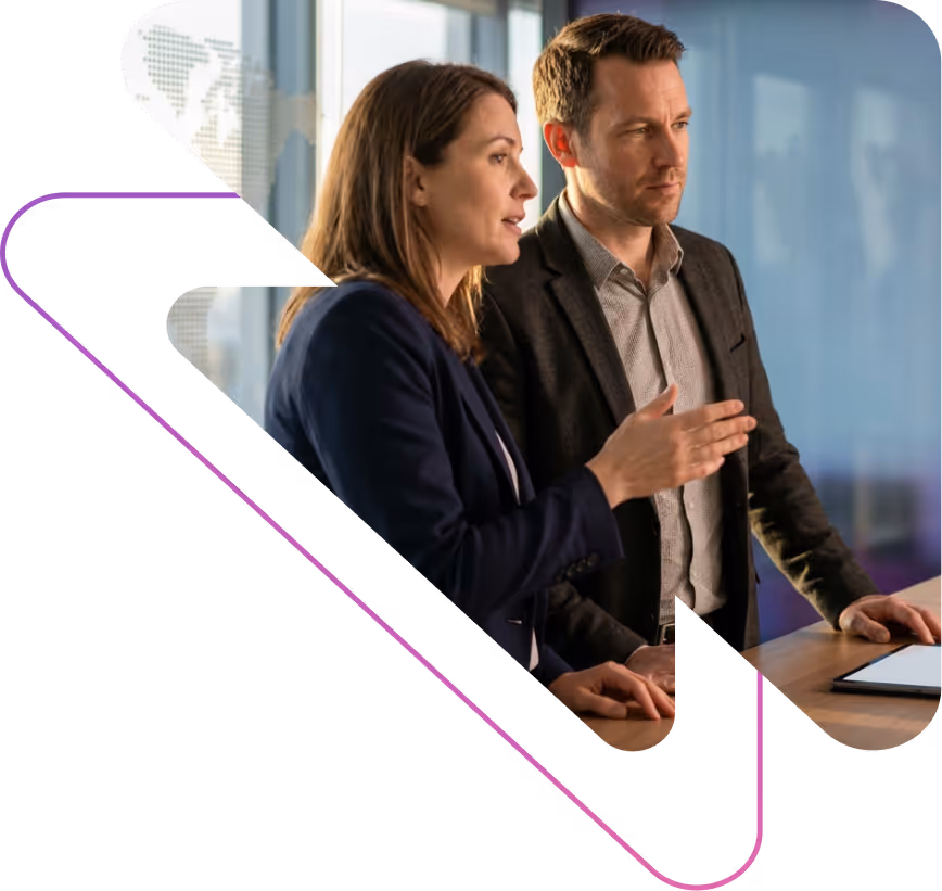 Two business professionals, a woman and a man, discussing while standing at a desk with a tablet in an office setting.