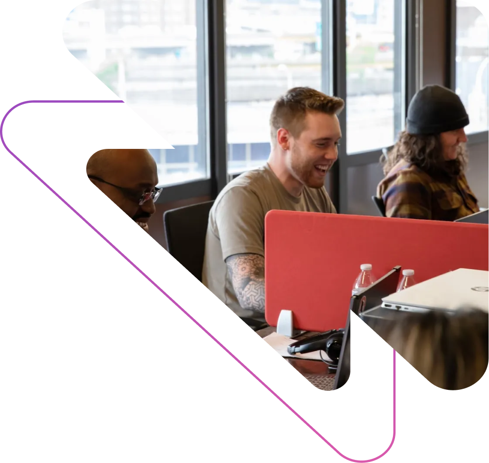 Three diverse coworkers laughing and working together at a desk with laptops in a bright office.