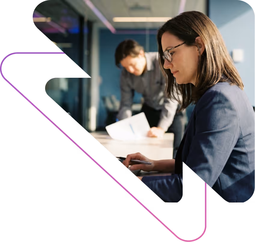 Woman with glasses in a navy blazer working on a laptop while a man in the background reviews papers in an office.