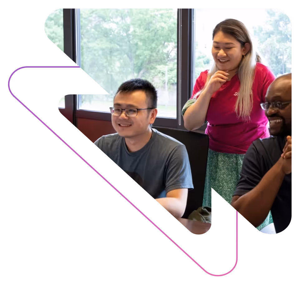 Three diverse colleagues smiling and collaborating in an office near a large window with trees outside.