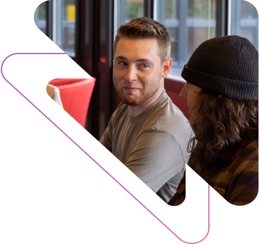 Two people sitting indoors, one person with short hair smiling at the other wearing a black beanie.