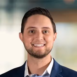 Smiling man with short dark hair, beard, and wearing a dark blazer over a white shirt against a blurred background.